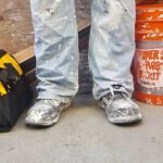 Photograph from the knees down of paint splattered jeans and boots next to a bucket and tool case